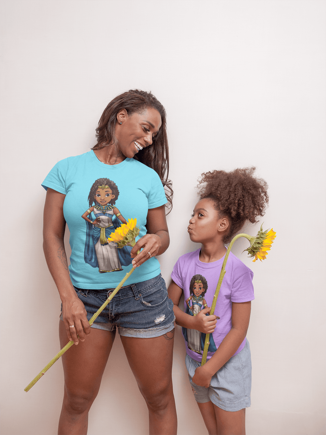 Black woman with her daughter both wearing Nefertiti T-Shirts. holding sunflowers. The mother's shirt is teal the daughter's is purple 