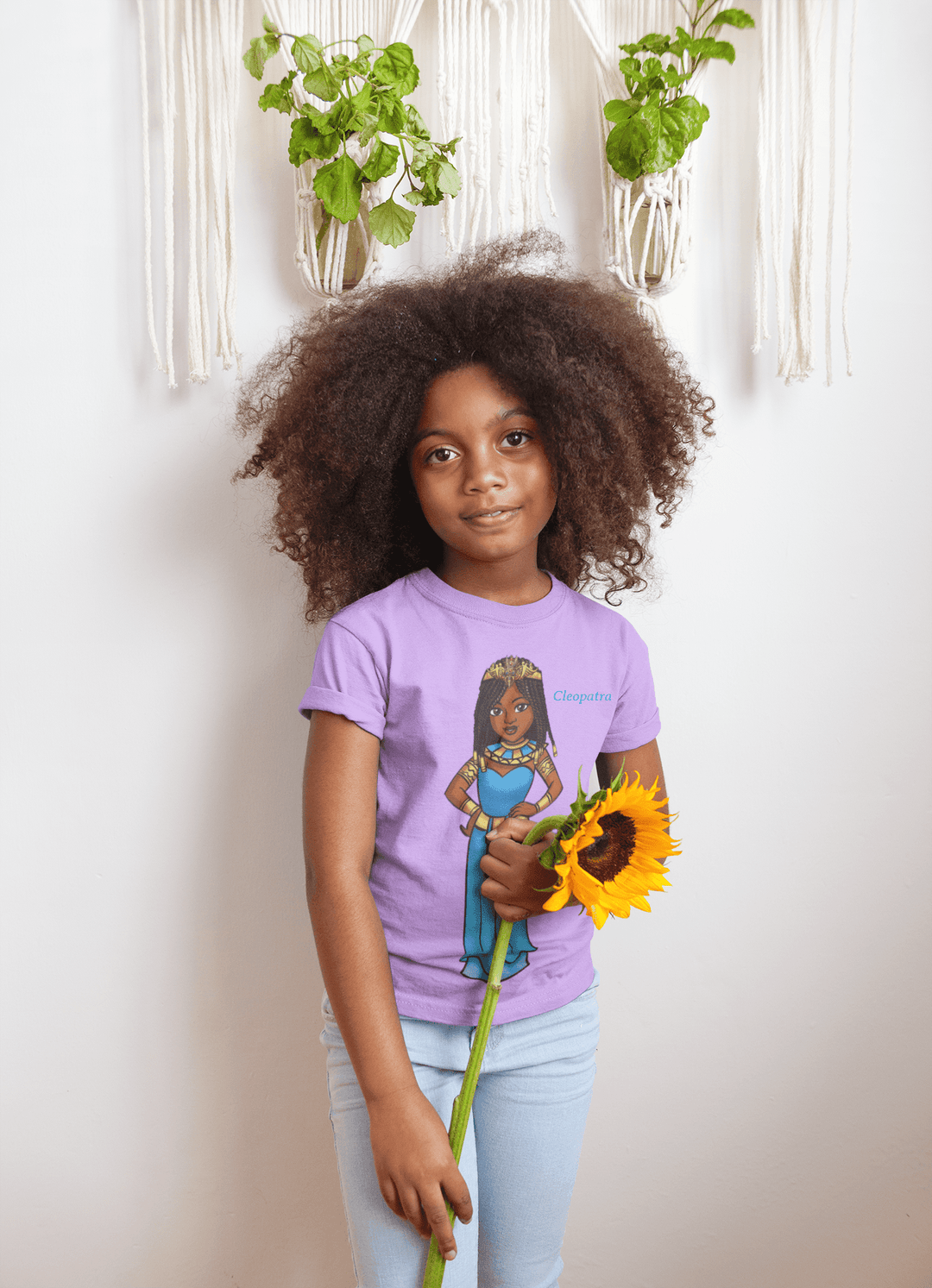Young Black girl wearing a lavender Cleopatra T-shirt from TL Johnson Stories, holding a sunflower. The shirts represent cultural empowerment fashion for children.